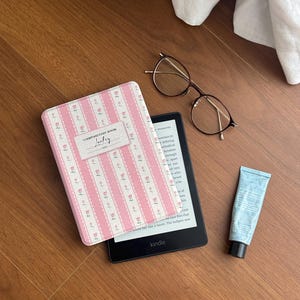 May include: A pink and white floral patterned Kindle cover with the text "Composition Book Juckey" rests on a wooden surface next to a Kindle e-reader, eyeglasses, and a tube of blue lotion.