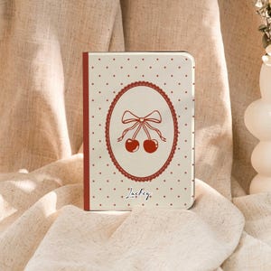 May include: A cream-colored notebook with a red border and a cherry design. The cover features a red oval with a bow and two cherries, surrounded by a dotted pattern. The word "Lucky" is written in cursive.