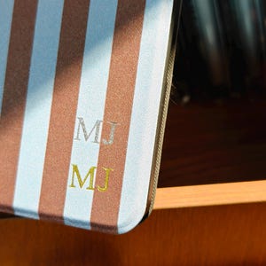 Puede incluir: Una cubierta de cuaderno a rayas verticales marrones y celeste claro. Las iniciales "MJ" est&aacute;n bordadas en hilo plateado y dorado. El cuaderno es parcialmente visible, descansando en un caj&oacute;n de madera.