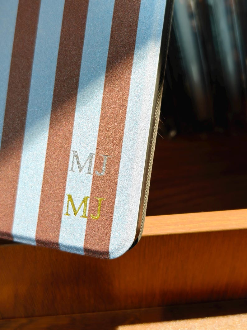 Puede incluir: Una cubierta de cuaderno a rayas verticales marrones y celeste claro. Las iniciales "MJ" est&aacute;n bordadas en hilo plateado y dorado. El cuaderno es parcialmente visible, descansando en un caj&oacute;n de madera.