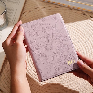 May include: A lavender passport holder with an embossed floral design. The cover features a detailed pattern of flowers and leaves. The initials "BO" are in gold on the lower right corner. The holder is held by hands.