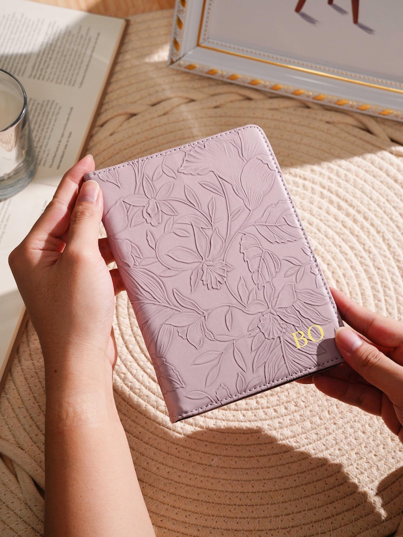 May include: A lavender passport holder with an embossed floral design. The cover features a detailed pattern of flowers and leaves. The initials "BO" are in gold on the lower right corner. The holder is held by hands.