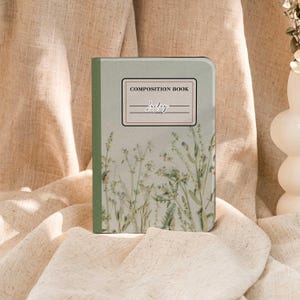May include: A composition book with a light green cover featuring a floral design. The cover includes the text "COMPOSITION BOOK" and the word "Lucky" in cursive. The book is rectangular and rests on a beige fabric surface.