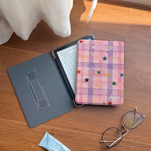 May include: An open gray e-reader case with a built-in stand next to a pink plaid e-reader cover decorated with colorful stars. A tube of blue lotion and a pair of round eyeglasses rest on a wooden surface.