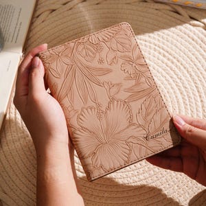 May include: A tan leather passport holder with an embossed floral design featuring a large hibiscus flower and leaves. The name "Camila" is written in script on the lower right corner. The holder is held open.