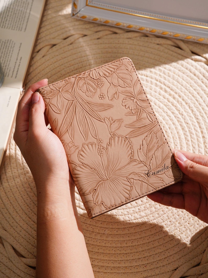 May include: A tan leather passport holder with an embossed floral design featuring a large hibiscus flower and leaves. The name "Camila" is written in script on the lower right corner. The holder is held open.