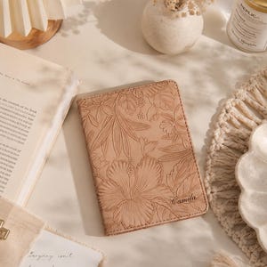 May include: A light brown passport holder with a floral design and the name "Camila" embossed on the front. The cover features a large hibiscus flower and other tropical leaves. The passport holder is on a light-colored surface.