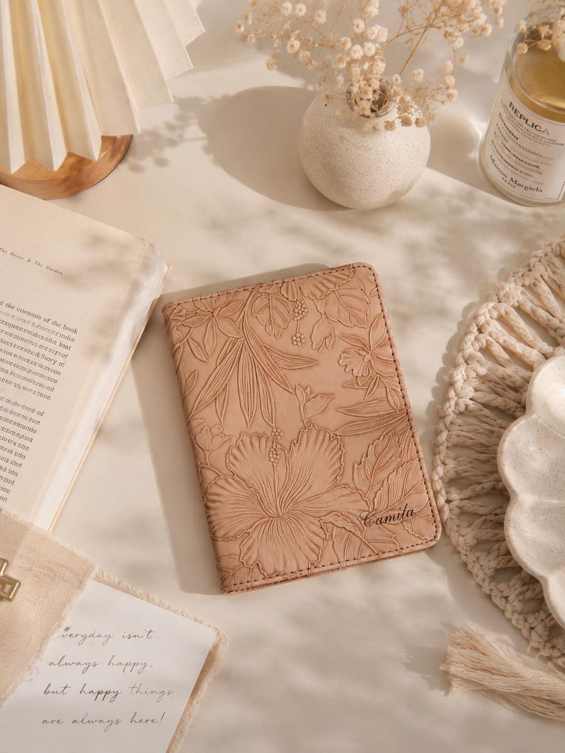 May include: A light brown passport holder with a floral design and the name "Camila" embossed on the front. The cover features a large hibiscus flower and other tropical leaves. The passport holder is on a light-colored surface.