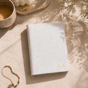 May include: A white passport holder with a floral embossed design and gold initials "KK". A gold bracelet with a small charm is next to the passport holder. A cup of coffee, a candle, and a bottle of perfume are also in the image.