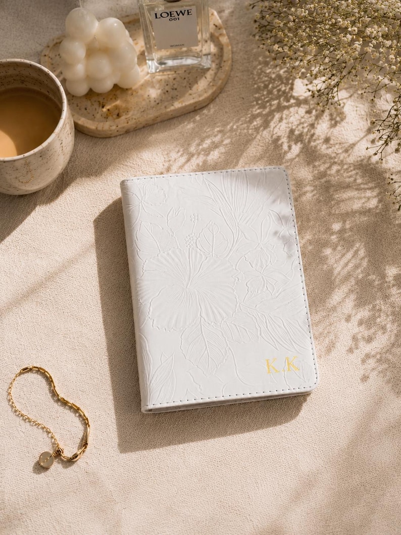 May include: A white passport holder with a floral embossed design and gold initials "KK". A gold bracelet with a small charm is next to the passport holder. A cup of coffee, a candle, and a bottle of perfume are also in the image.