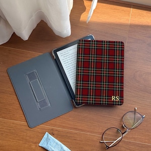 May include: A red, green, and black plaid e-reader case with the gold initials RS on the front cover. The case is open, revealing a gray interior with a hand strap, and is accompanied by a small light blue tube and round-framed glasses.