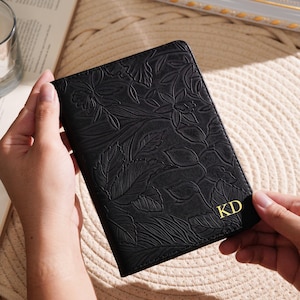 May include: Black passport holder with embossed floral design. The cover features gold-colored initials "KD" in the lower right corner. The passport holder is held by a person.