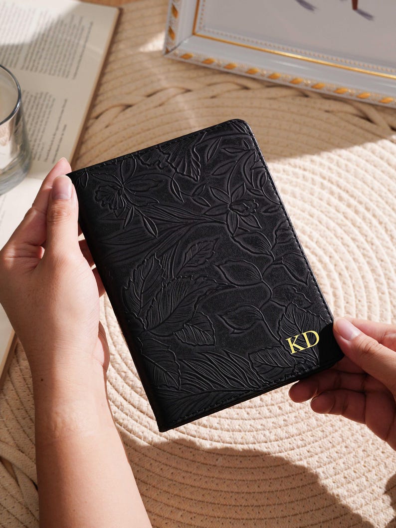 May include: Black passport holder with embossed floral design. The cover features gold-colored initials "KD" in the lower right corner. The passport holder is held by a person.