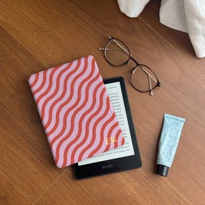May include: A Kindle e-reader with a pink and red wavy patterned cover, next to a pair of eyeglasses and a tube of blue lotion. The e-reader displays text and the word "kindle". The cover has the initials "ER".
