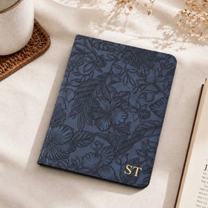 May include: A rectangular navy blue passport holder featuring an embossed floral pattern across the entire surface. The bottom right corner displays the gold-colored capital letters ST. The cover has rounded corners and a smooth, matte finish.