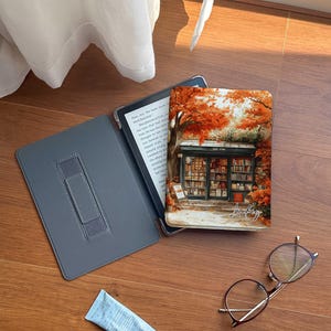 May include: A gray e-reader with a black cover and a brown leather strap. The e-reader is open to a page with text. The e-reader is sitting on a wooden table next to a book with a colorful cover featuring a bookstore with autumn leaves. The book cover also has the word "Lucky" written on it. There are also a pair of glasses and a tube of hand cream on the table.