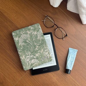May include: A black Kindle e-reader with a floral patterned case. The e-reader is open to a page with text. A tube of hand cream and a pair of spectacles are also in the image.