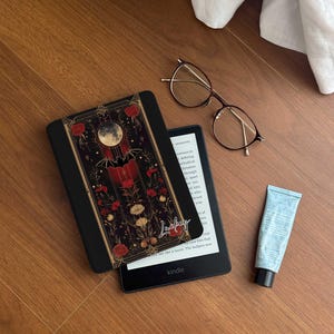 May include: A black Kindle e-reader with a floral and moon design case. The case has a gold trim and a bat silhouette. The text "Jackey" is written on the case. The Kindle is on a wooden surface with a pair of glasses and a tube of lotion.