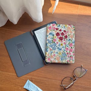 以下が含まれることがあります： 木製の表面に、グレーの電子書籍リーダーと花柄のケース、ハンドクリームのチューブ、丸い眼鏡が置かれています。