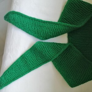 May include: A knitted green scarf, displayed on a white surface. The scarf is made of a thick yarn, with a visible knit pattern. The scarf is folded in a way that shows its length and width. The color is a vibrant, solid green.