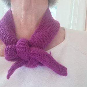 May include: A knitted purple scarf tied in a knot. The scarf is made of a thick yarn and is wrapped around the neck. The scarf is a solid color and has a textured appearance.
