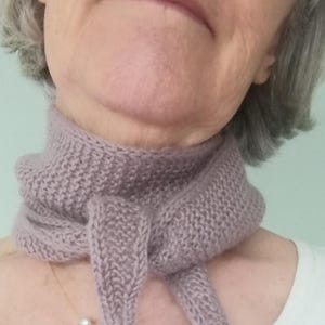 May include: A knitted lavender scarf with a textured pattern. The scarf is wrapped around the neck and tied in a knot. The scarf is made of a soft, fuzzy material.