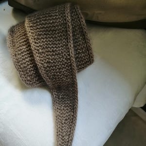 May include: A hand-knitted scarf in shades of brown, with a textured appearance. The scarf is folded and draped, showcasing its length and the detailed stitch work. The neutral color palette makes it a versatile accessory.