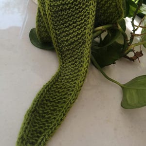 May include: A knitted green scarf, folded and draped. The scarf is a solid, medium green color with a textured, ribbed pattern. The scarf is made of a soft yarn and appears to be handmade. The scarf is shown on a white surface with green leaves in the background.