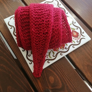 May include: A folded, hand-knitted scarf in a vibrant red hue. The scarf is made of thick yarn, with a textured appearance. It is resting on a decorative tile with floral patterns and a wooden surface.