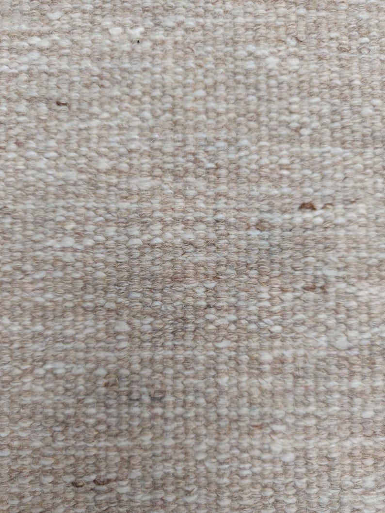K&ouml;nnte beinhalten: Ein dicht gewebter Textilstoff mit einer dichten, noppigen Struktur in einer neutralen Farbpalette. Das Material zeigt eine Mischung aus hellbeigen, hellbraunen und cremewei&szlig;en Fasern, die ein gleichm&auml;&szlig;iges Erscheinungsbild auf der Oberfl&auml;che erzeugen.
