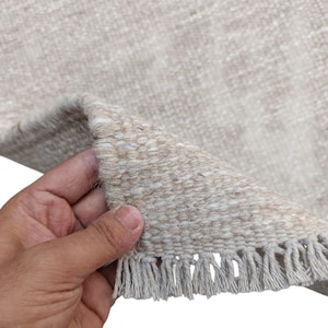 K&ouml;nnte beinhalten: Ein gewebter Teppich in neutralen Beige- und Off-White-T&ouml;nen mit einer dicken, strukturierten Webart. Der Teppich hat eine umgeschlagene Ecke, die die dichte Struktur zeigt, und ist mit einer Reihe kurzer, gerader Fransen am Rand versehen.