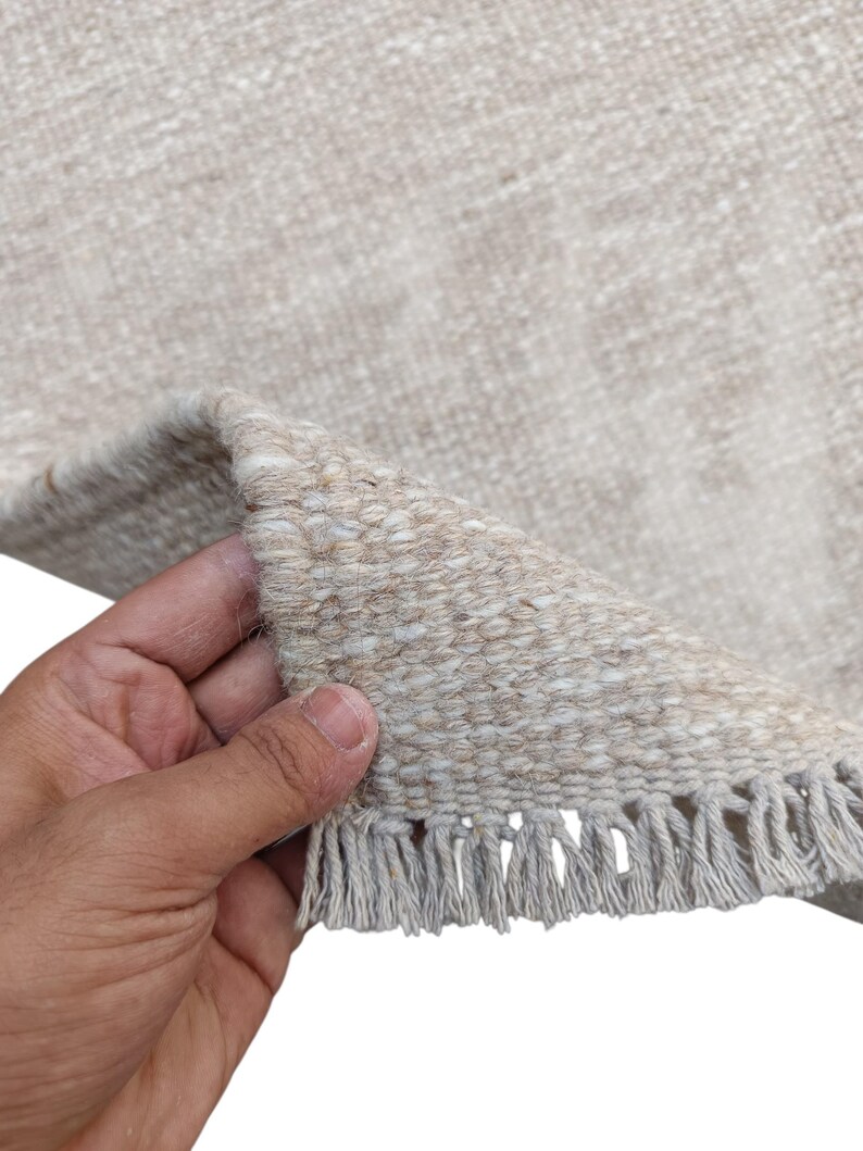 K&ouml;nnte beinhalten: Ein gewebter Teppich in neutralen Beige- und Off-White-T&ouml;nen mit einer dicken, strukturierten Webart. Der Teppich hat eine umgeschlagene Ecke, die die dichte Struktur zeigt, und ist mit einer Reihe kurzer, gerader Fransen am Rand versehen.