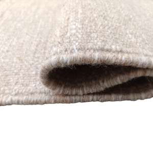 K&ouml;nnte beinhalten: Ein rechteckiger Webteppich mit einer strukturierten Oberfl&auml;che in Beige-, Hellbraun- und Cremet&ouml;nen. Der Teppich ist an einem Ende umgeschlagen und zeigt die dicke, dichte Konstruktion der Fasern sowie die sauber eingefassten Kanten.