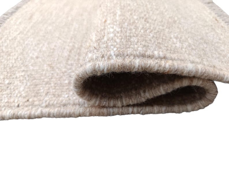 K&ouml;nnte beinhalten: Ein rechteckiger Webteppich mit einer strukturierten Oberfl&auml;che in Beige-, Hellbraun- und Cremet&ouml;nen. Der Teppich ist an einem Ende umgeschlagen und zeigt die dicke, dichte Konstruktion der Fasern sowie die sauber eingefassten Kanten.