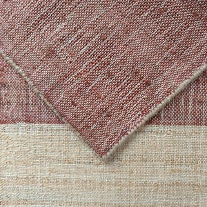 May include: A woven fabric swatch featuring a textured, coarse weave with horizontal stripes in muted terracotta red and off-white. The fabric is folded at an angle, revealing the underside of the terracotta section with its distinct, thick, fibrous weave.