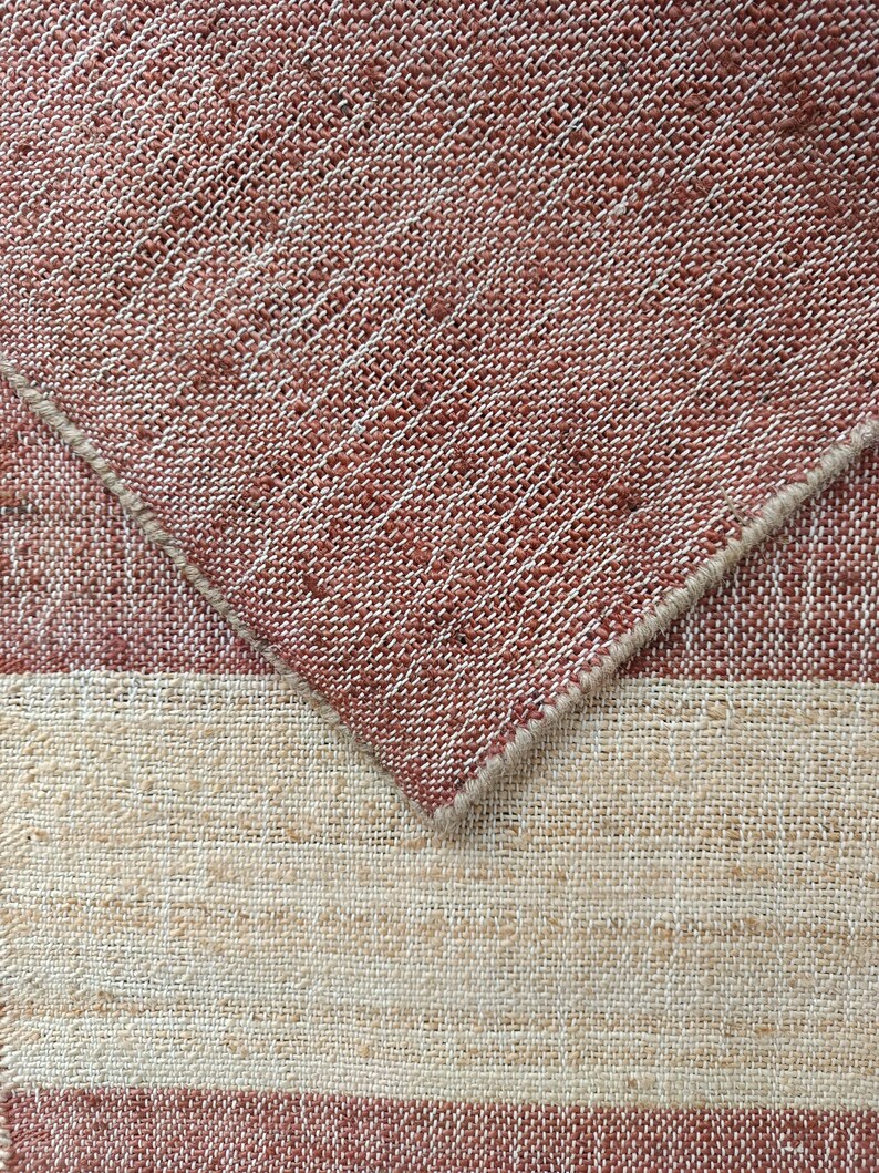 May include: A woven fabric swatch featuring a textured, coarse weave with horizontal stripes in muted terracotta red and off-white. The fabric is folded at an angle, revealing the underside of the terracotta section with its distinct, thick, fibrous weave.