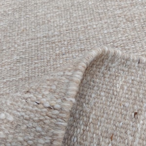 K&ouml;nnte beinhalten: Ein gewebter Teppich in neutralen Beige- und Off-White-T&ouml;nen mit einer dicken, strukturierten Garnkonstruktion. Der Stoff zeigt ein sichtbares Flechtmuster mit einer umgeschlagenen Ecke, die die Unterseite freigibt, und einer Fransenborte.