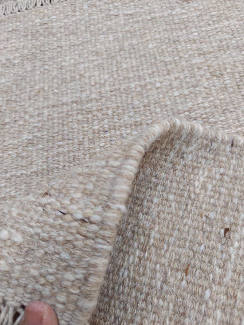 K&ouml;nnte beinhalten: Ein gewebter Teppich in neutralen Beige- und Off-White-T&ouml;nen mit einer dicken, strukturierten Garnkonstruktion. Der Stoff zeigt ein sichtbares Flechtmuster mit einer umgeschlagenen Ecke, die die Unterseite freigibt, und einer Fransenborte.