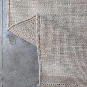 Pu&ograve; includere: Un tappeto intrecciato in una tonalit&agrave; beige neutra con una struttura a pelo riccio spesso e testurizzato. Il tappeto &egrave; ripiegato su un angolo per rivelare il retro, e il bordo &egrave; rifinito con corte frange annodate lungo il lato inferiore.
