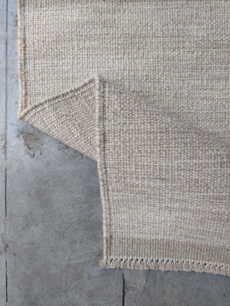 Pu&ograve; includere: Un tappeto intrecciato in una tonalit&agrave; beige neutra con una struttura a pelo riccio spesso e testurizzato. Il tappeto &egrave; ripiegato su un angolo per rivelare il retro, e il bordo &egrave; rifinito con corte frange annodate lungo il lato inferiore.