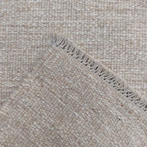 K&ouml;nnte beinhalten: Ein gewebter Teppich in neutralen Beige- und Cremet&ouml;nen mit einer strukturierten, noppigen Oberfl&auml;che. Der Teppich ist an einer Ecke umgeschlagen und zeigt eine Kante mit kurzen, dichten Fransen entlang der diagonalen Falzlinie des Umschlags.
