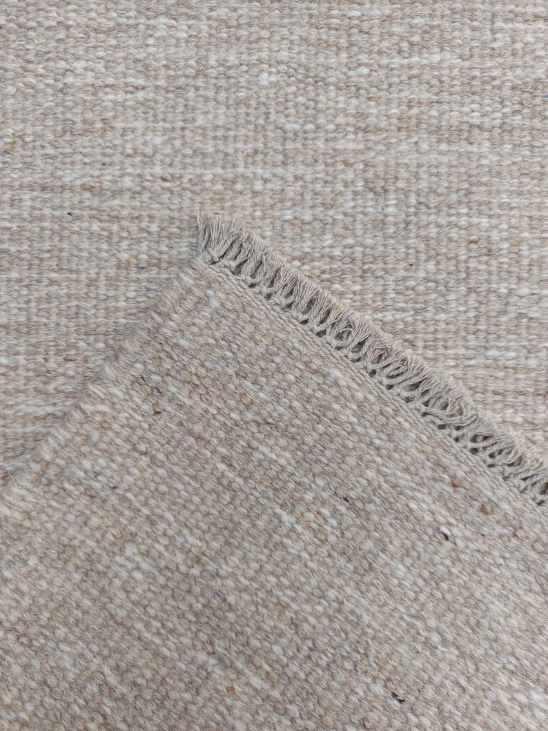 K&ouml;nnte beinhalten: Ein gewebter Teppich in neutralen Beige- und Cremet&ouml;nen mit einer strukturierten, noppigen Oberfl&auml;che. Der Teppich ist an einer Ecke umgeschlagen und zeigt eine Kante mit kurzen, dichten Fransen entlang der diagonalen Falzlinie des Umschlags.