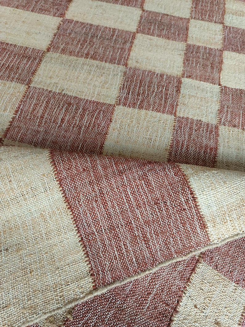 May include: A woven textile fabric featuring a repeating checkerboard pattern in alternating squares of natural beige and terracotta red. The material has a visible coarse texture with distinct stitching lines defining each individual square.