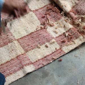 May include: A woven rug featuring a checkered pattern of alternating cream and terracotta-colored squares. The fabric has a coarse, fibrous texture with loose, frayed fibers scattered across the surface and along the stitched seams.