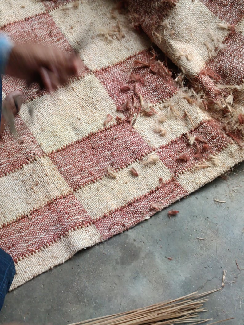 May include: A woven rug featuring a checkered pattern of alternating cream and terracotta-colored squares. The fabric has a coarse, fibrous texture with loose, frayed fibers scattered across the surface and along the stitched seams.