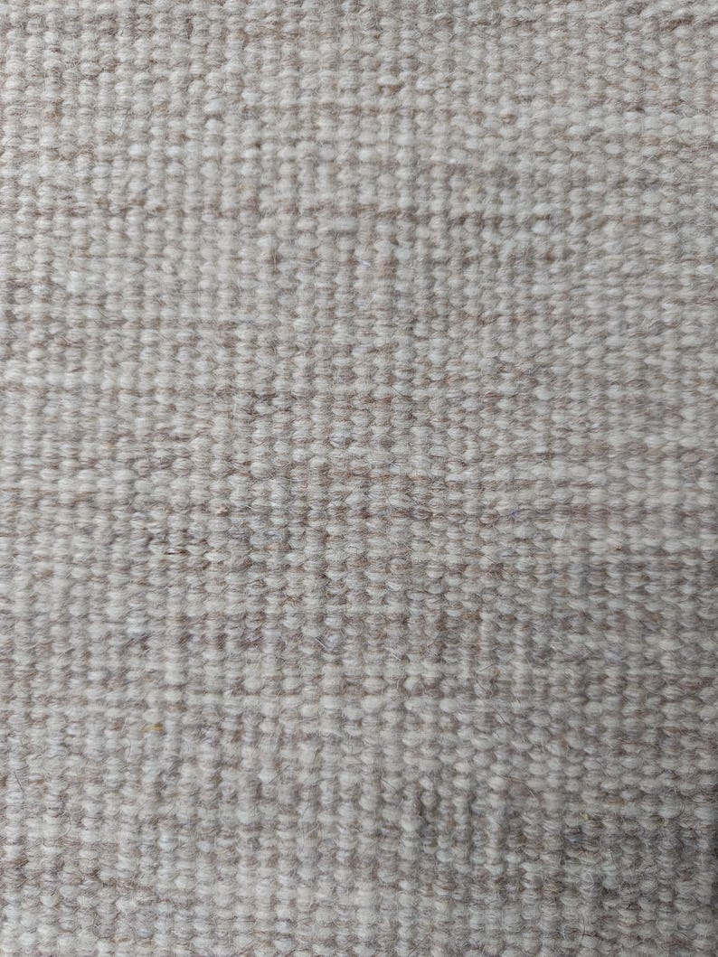 Pu&ograve; includere: Una vista ravvicinata di un tessuto intrecciato caratterizzato da una densa struttura a ricciolo. Il tessuto presenta una palette di colori neutri, che fonde fibre beige tenue e bianco sporco in un motivo a griglia costante e ripetitivo.