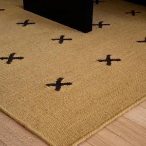 Puede incluir: Una alfombra rectangular con una superficie texturizada de color tostado y un patrón repetitivo de símbolos de cruz de color marrón oscuro distribuidos por la tela. La alfombra tiene un borde acabado y un tejido consistente en toda su superficie.