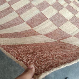 May include: A woven area rug featuring a repeating checkered pattern in alternating squares of terracotta orange and natural beige. The rug has a textured, fibrous surface with a visible weave and a finished edge along the perimeter of the fabric.