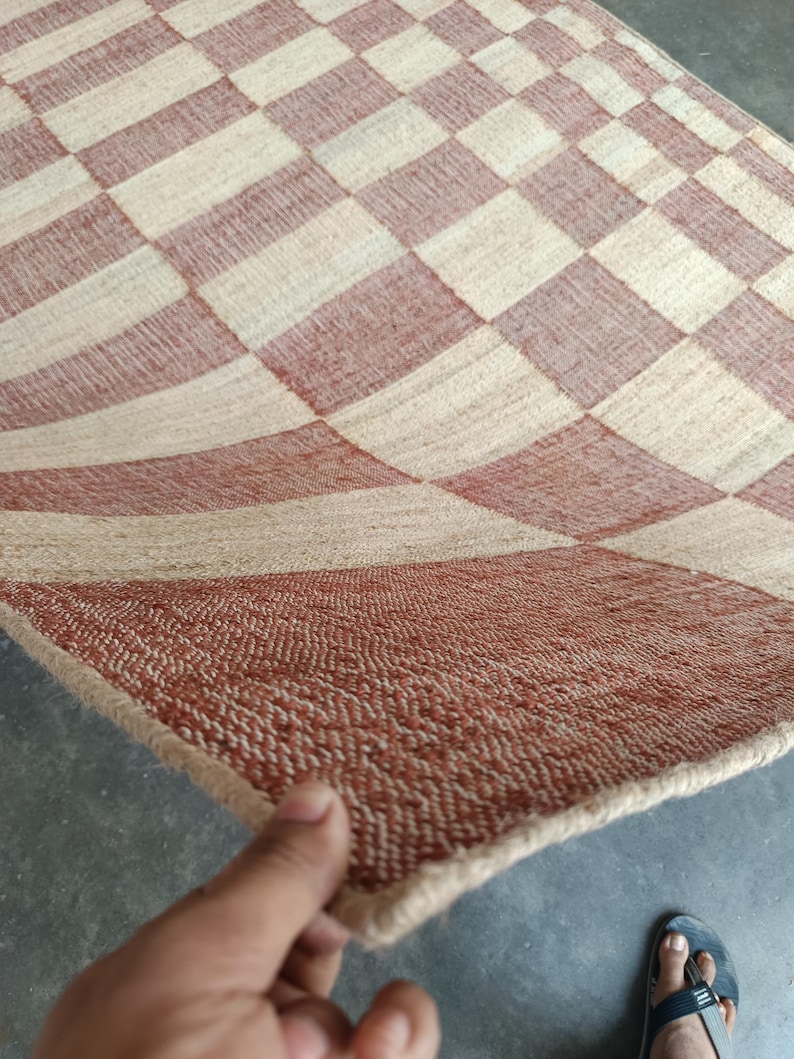 May include: A woven area rug featuring a repeating checkered pattern in alternating squares of terracotta orange and natural beige. The rug has a textured, fibrous surface with a visible weave and a finished edge along the perimeter of the fabric.