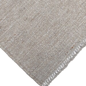 K&ouml;nnte beinhalten: Ein rechteckiger Webteppich in einer strukturierten, neutralen Farbpalette aus Beige und Off-White. Der Stoff zeigt ein dichtes, geschlungenes Webmuster mit dezenten braunen Sprenkeln. Eine Kante ist mit einer kurzen, geraden Franse versehen.